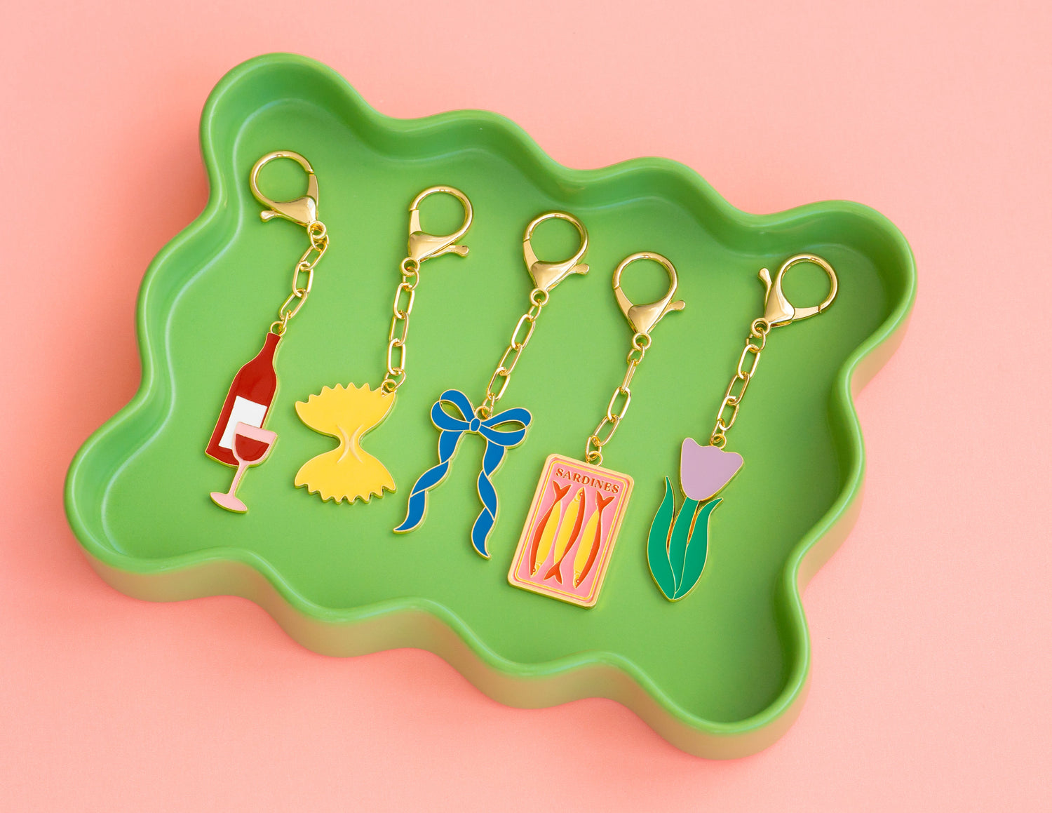 Five keychains with different enamel designs lay in a green, wavy tray on a pink background.