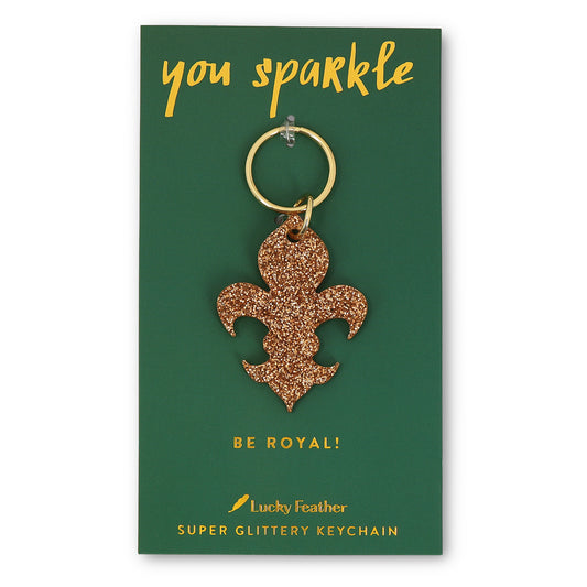A fleur-de-lis shaped gold glitter keychain on a green card that says 'you sparkle' and 'be royal!'.