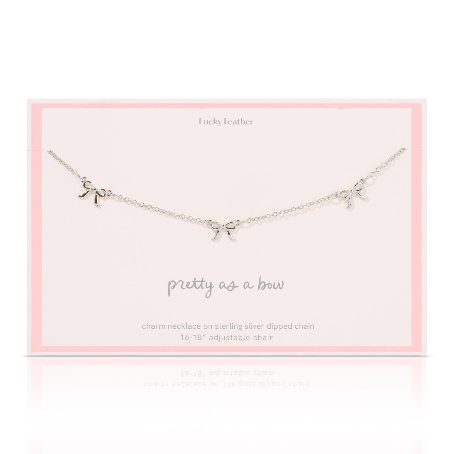 A silver-colored necklace with three bow charms lies on a pale pink card from 'Lucky Feather'.