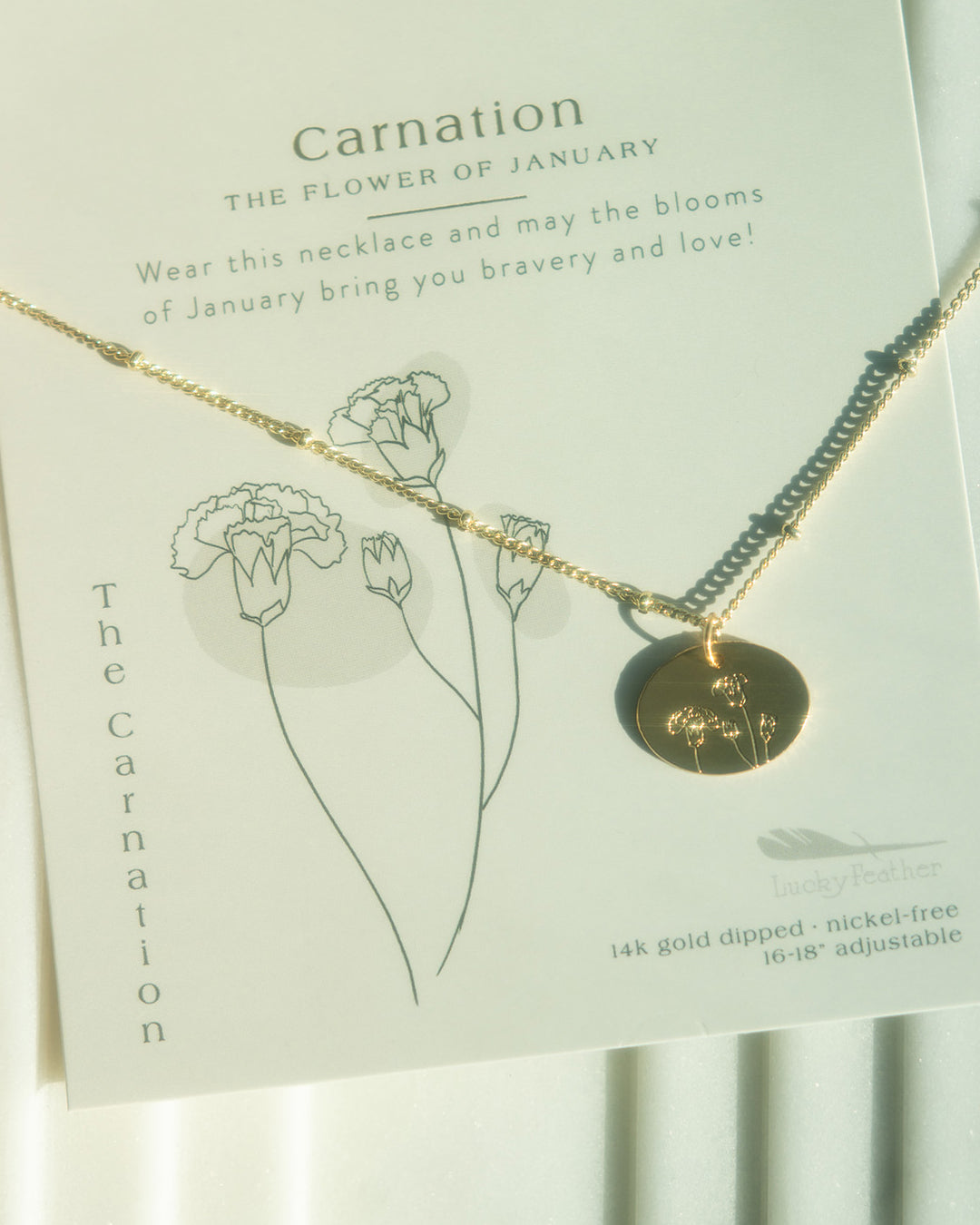A gold necklace with a flower-engraved pendant displayed on a card with carnation illustrations and text.