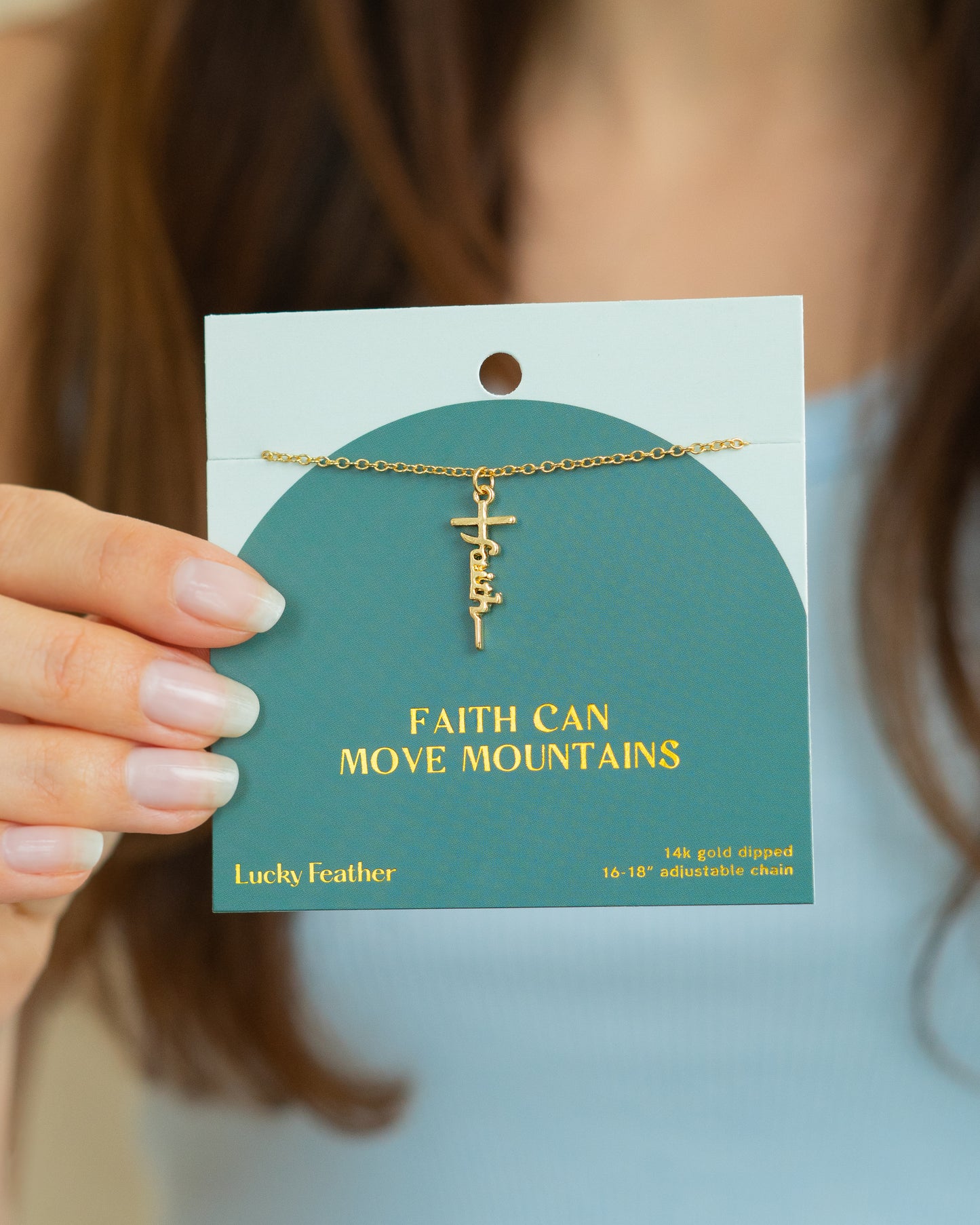 A person holds a teal card displaying a gold 'Faith' necklace and the words 'Faith Can Move Mountains'.