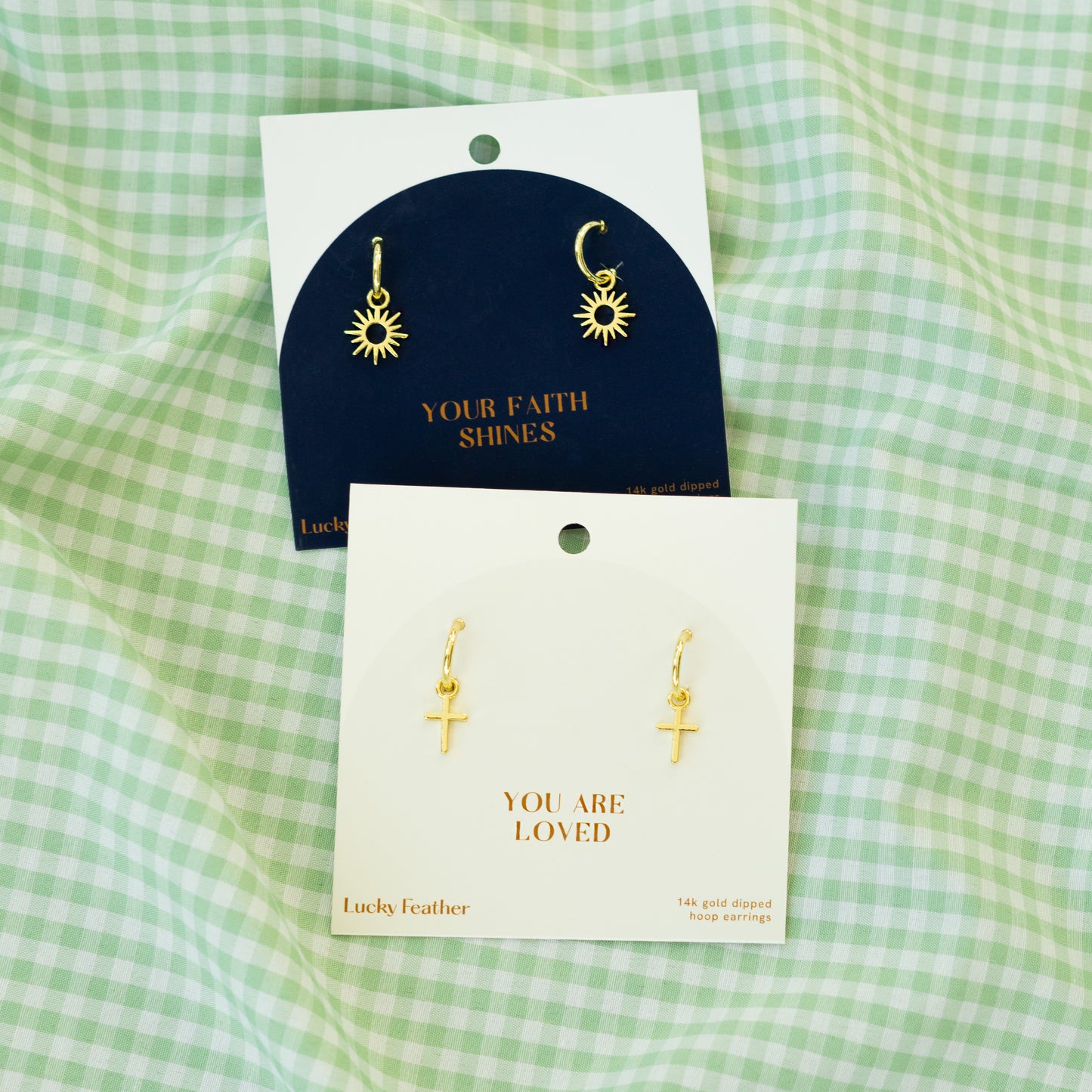 Two cards of earrings, one sun-shaped and the other cross-shaped, sit on a green-and-white checkered cloth.
