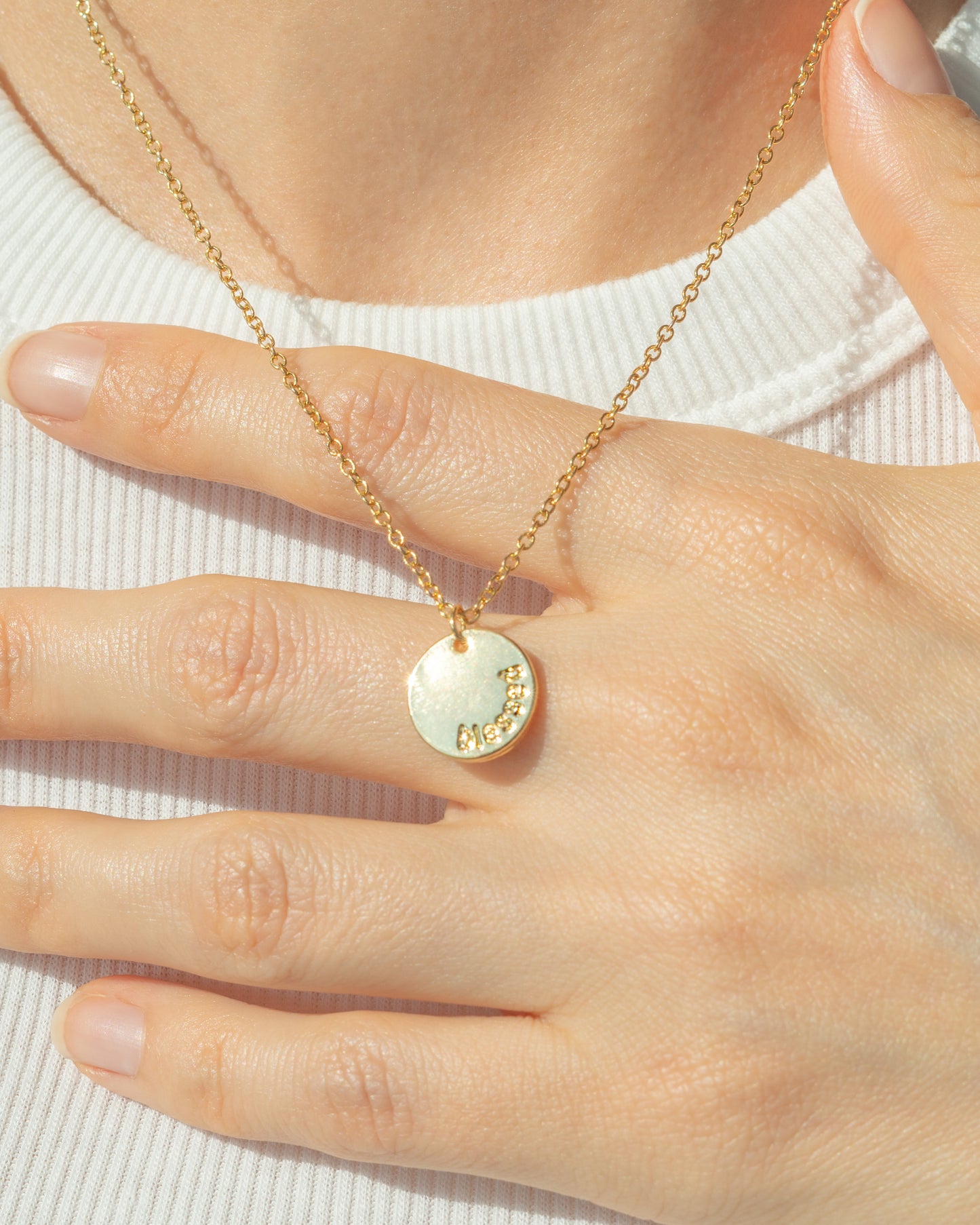 A gold necklace with a circular pendant is displayed on a person wearing a white shirt, with their hand resting on the pendant.