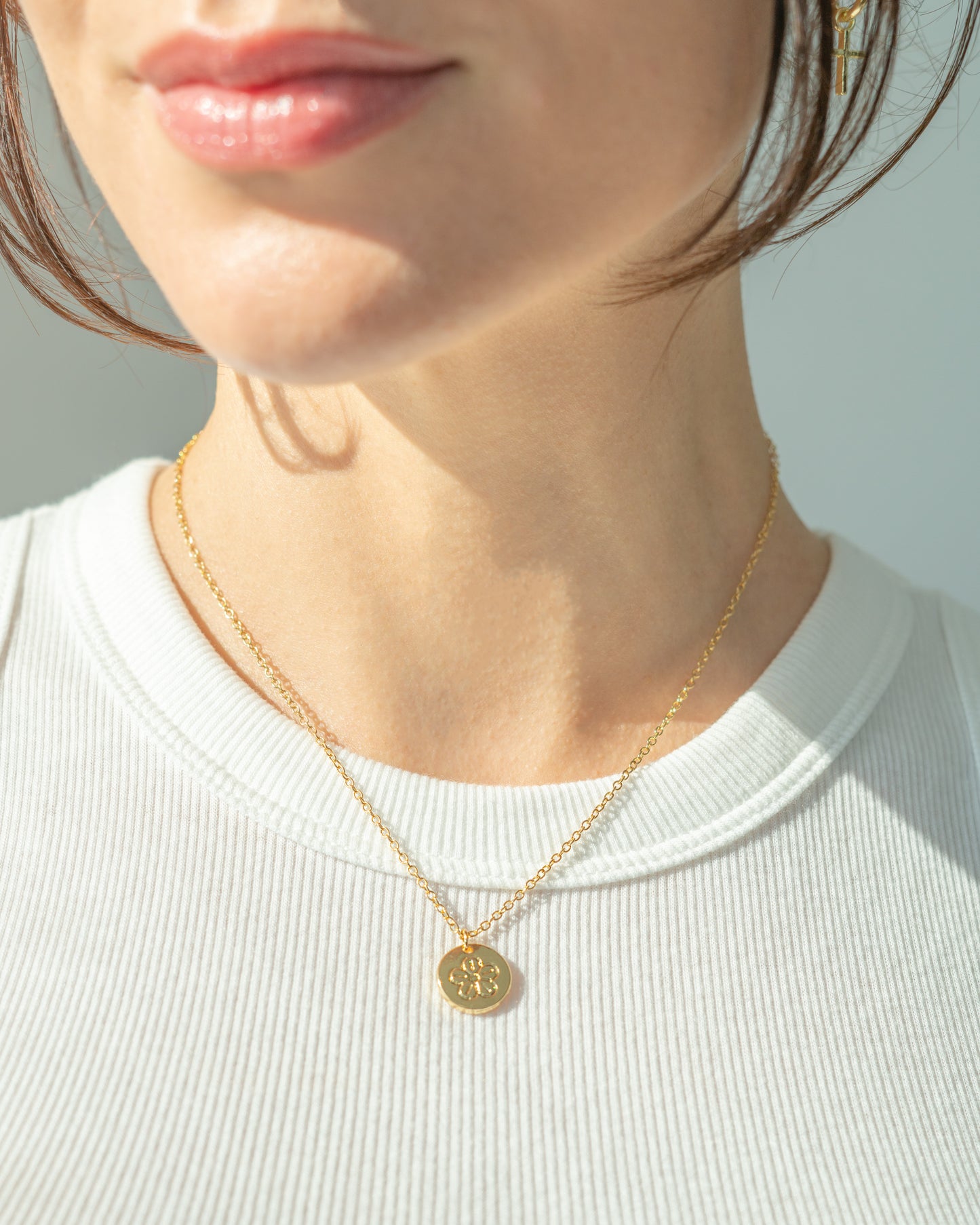 A woman wears a gold necklace with a floral pendant and a white ribbed shirt.