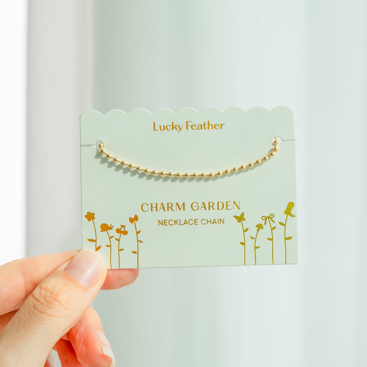 A hand holds a 'Lucky Feather Charm Garden Necklace Chain' product card, with a golden ball chain necklace attached.