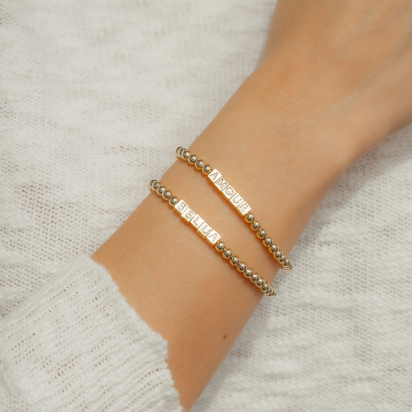 A wrist with two gold-colored beaded bracelets that spell out 'BELLA' and 'AMOUR' is pictured against a light-colored knitted background.