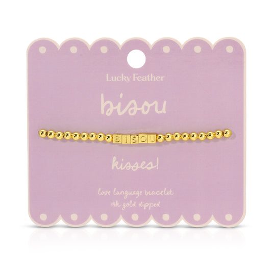A gold beaded bracelet with the word 'bisou' spelled out in square letter beads is displayed on a lavender card.