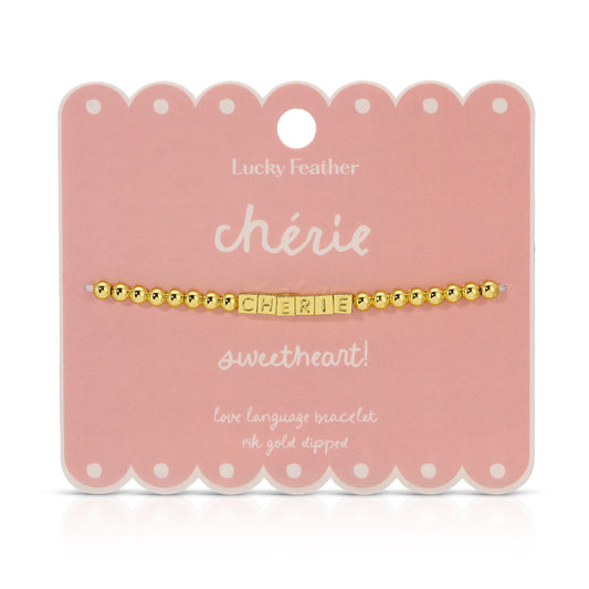 A 'chérie' bracelet with gold beads and lettered blocks is displayed on a scalloped pink card by Lucky Feather.