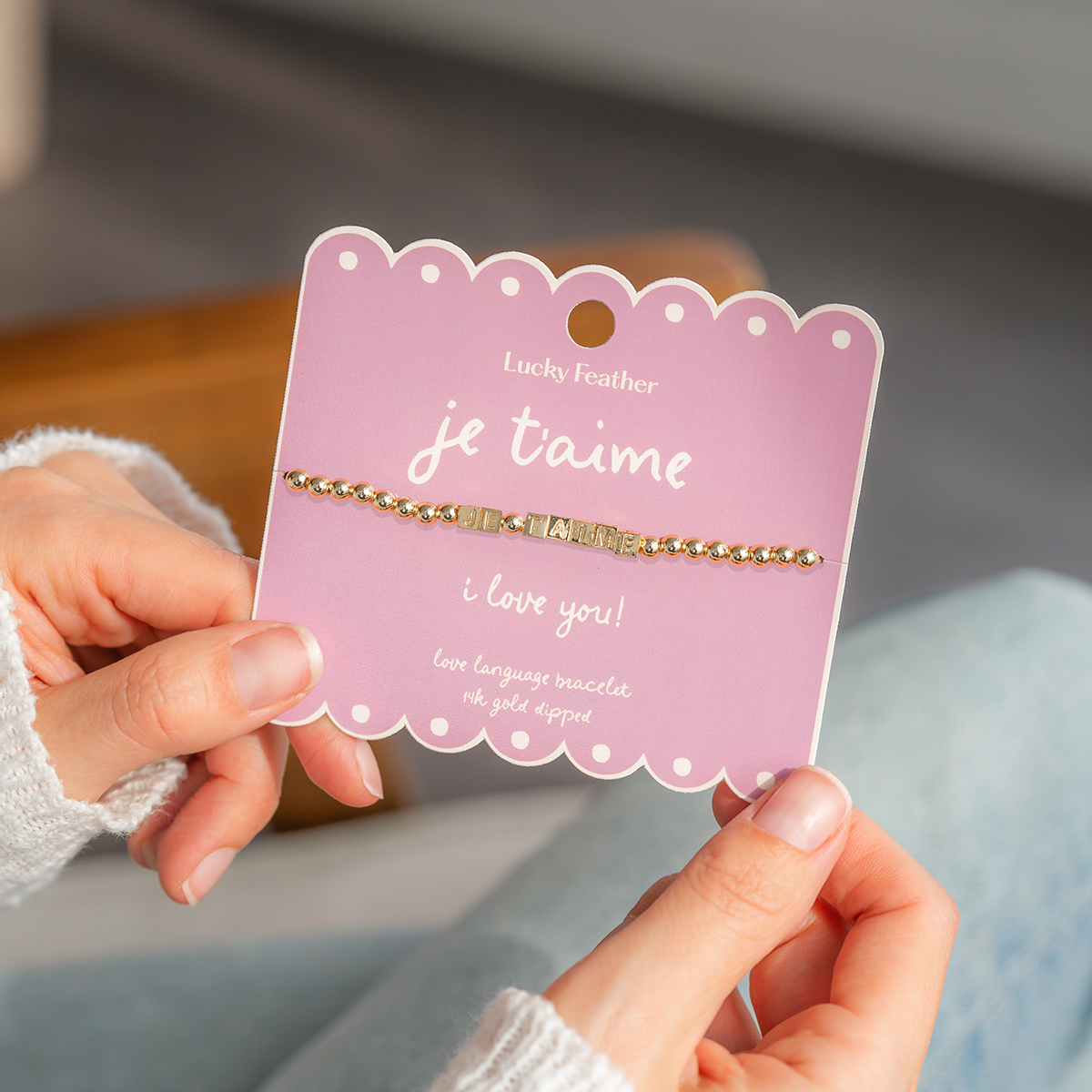 A person holds a 'Lucky Feather' card with a beaded bracelet that spells out 'JE T'AIME' and 'I love you!'.