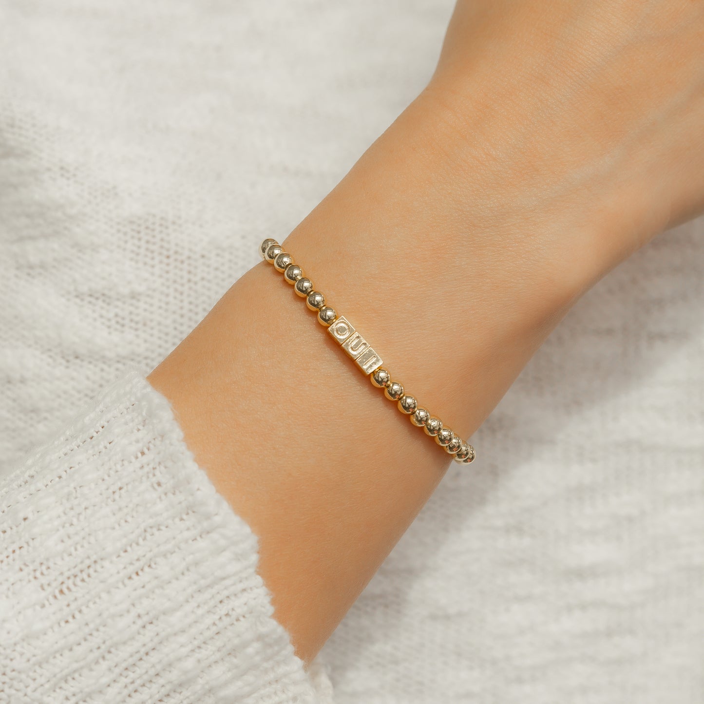 A gold beaded bracelet with a rectangular centerpiece bearing lettering is on a person's wrist, against a white sweater.