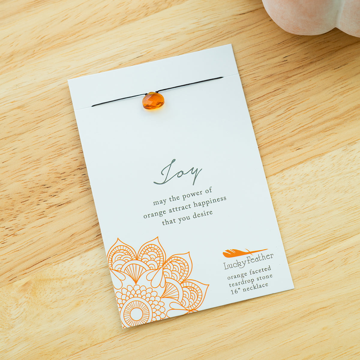 An orange teardrop stone necklace on a white card with the text "Joy, may the power of orange attract happiness that you desire" and the brand name "Lucky Feather".