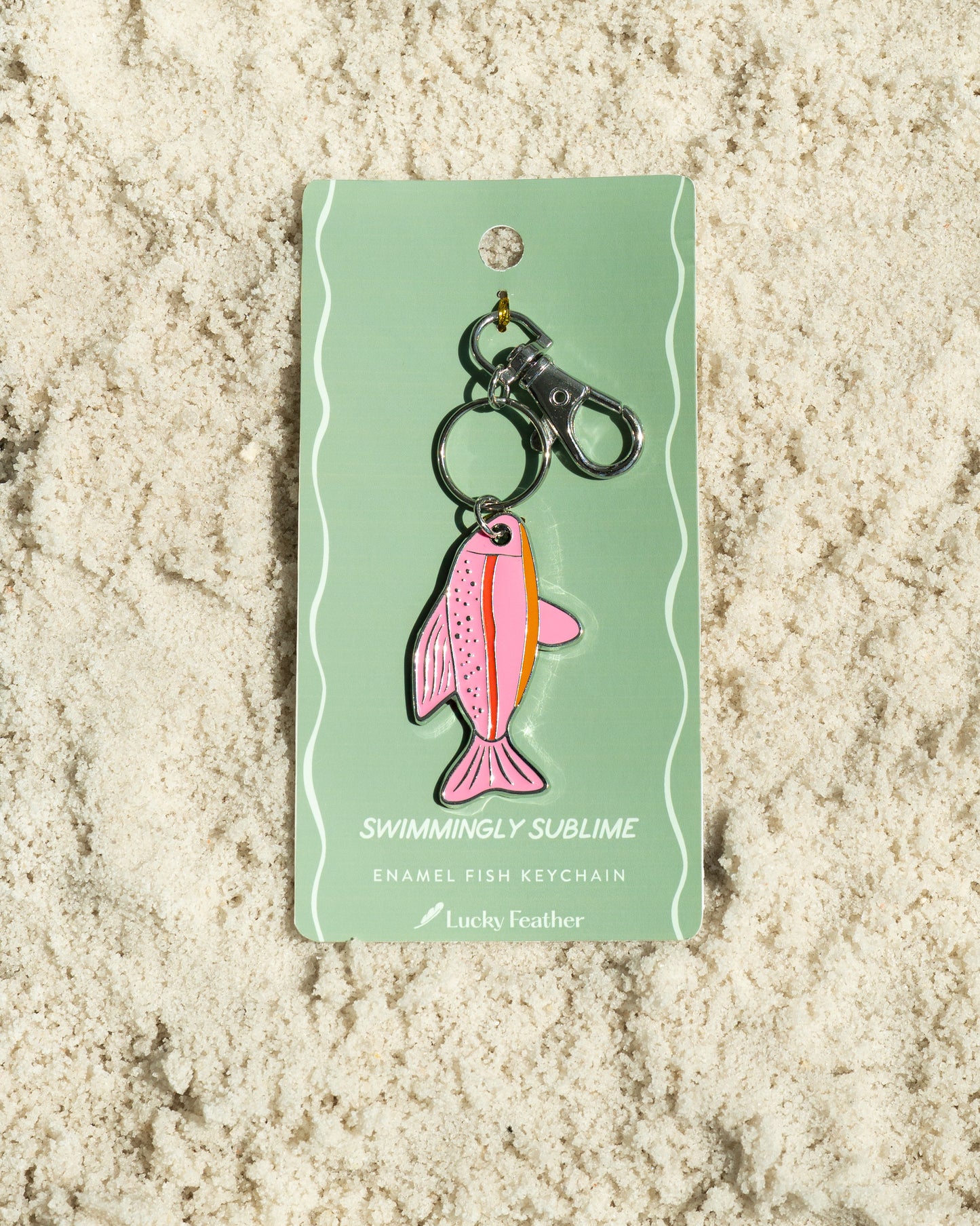A pink enamel fish keychain from 'Lucky Feather' is displayed on a light green card on a bed of light-colored sand.