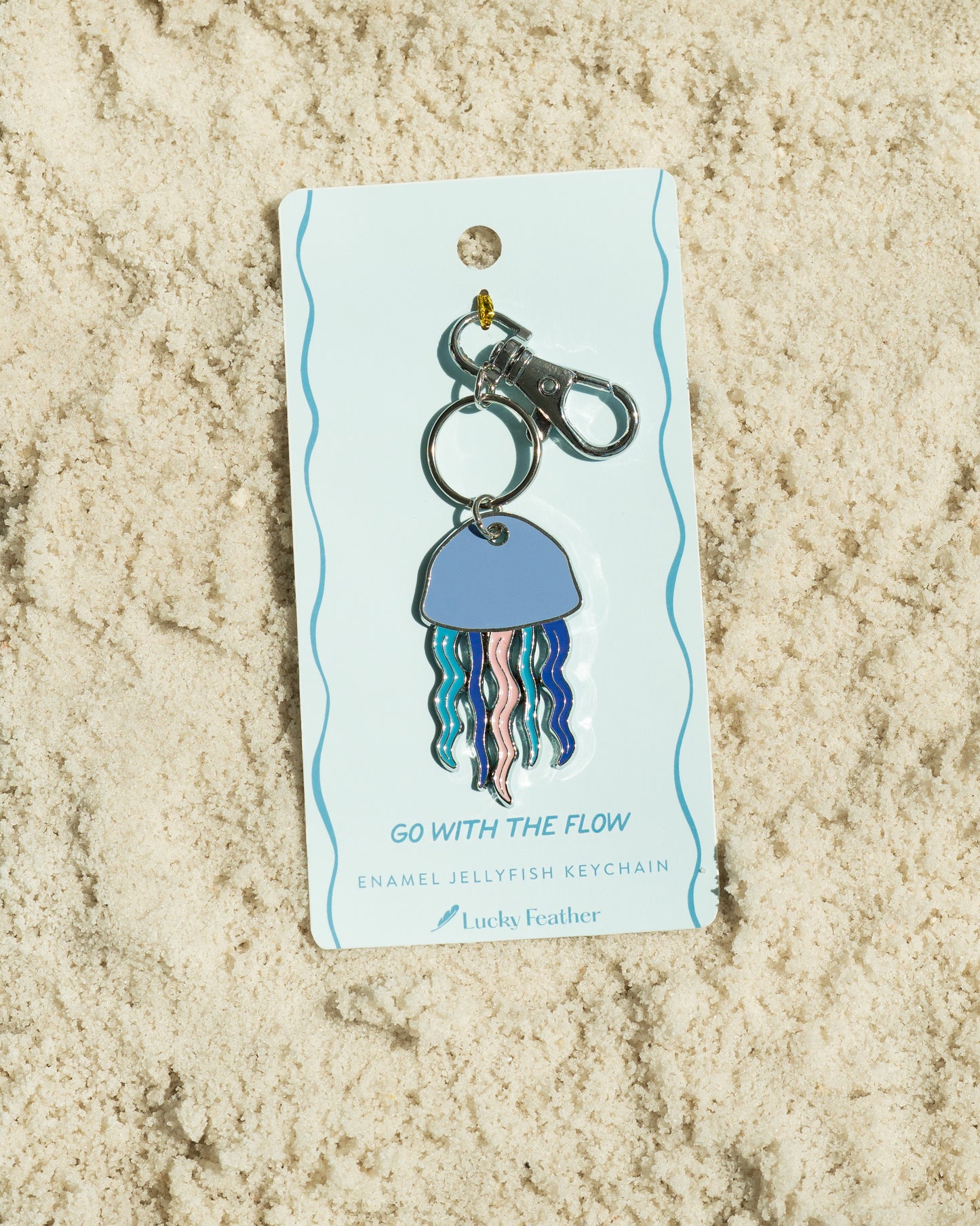 A 'Lucky Feather' brand enamel jellyfish keychain on its display card, resting on sand.