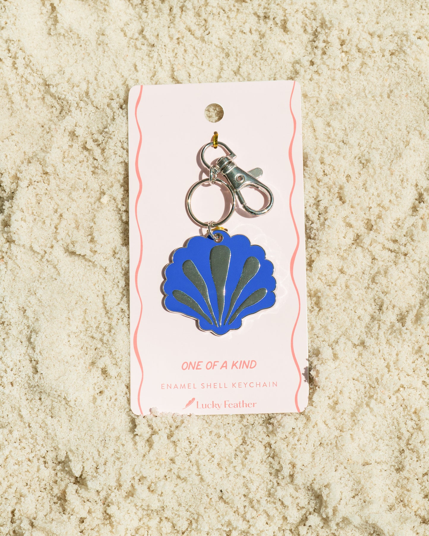 A blue and silver enamel seashell keychain attached to a pink display card with the words 'ONE OF A KIND' and 'ENAMEL SHELL KEYCHAIN' on sand.