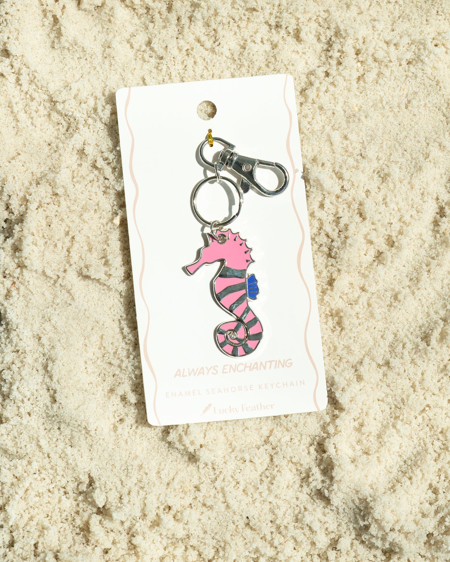 A seahorse keychain on a tag is displayed on a bed of sand.