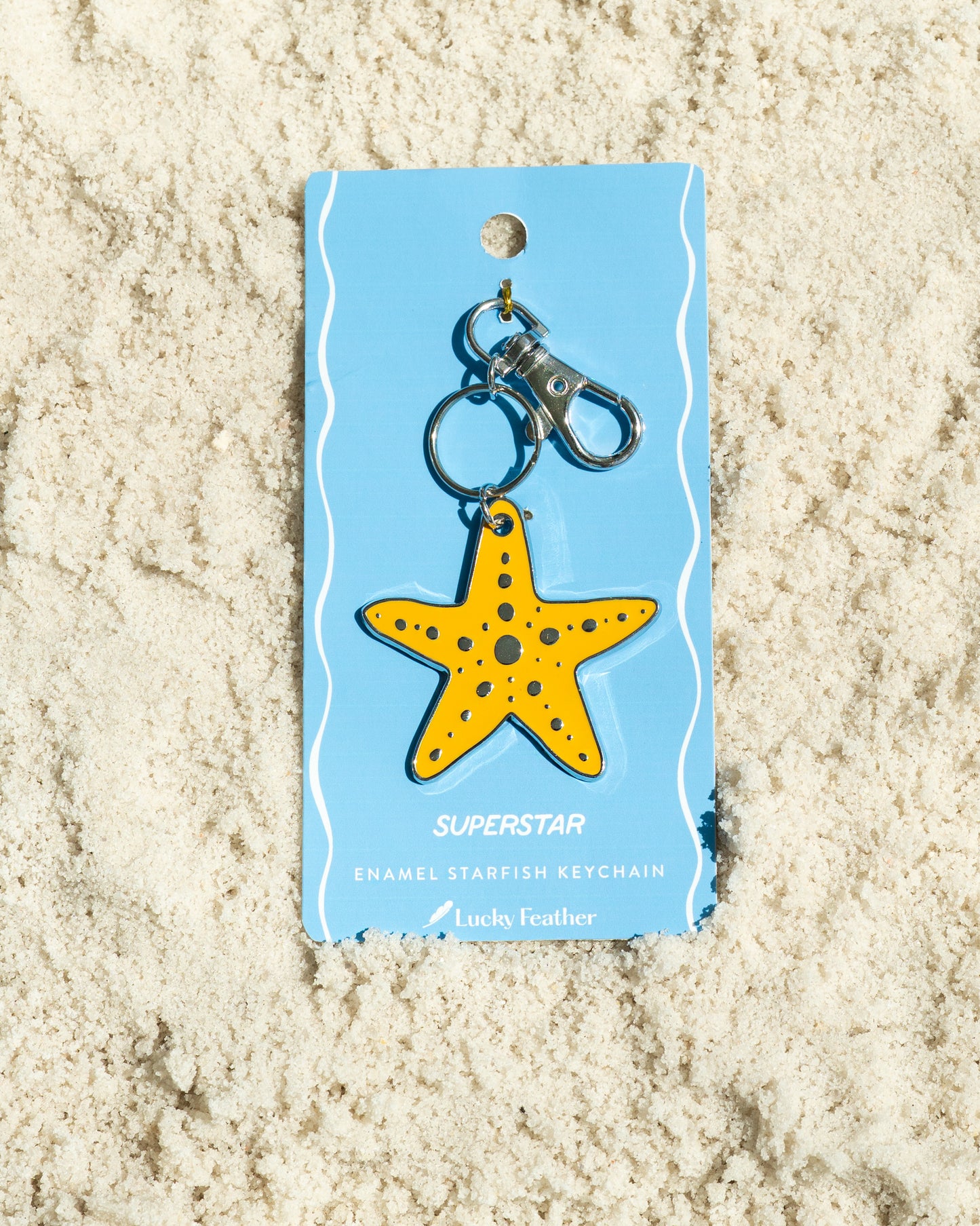 A starfish keychain from Lucky Feather is displayed on a sand-like surface.