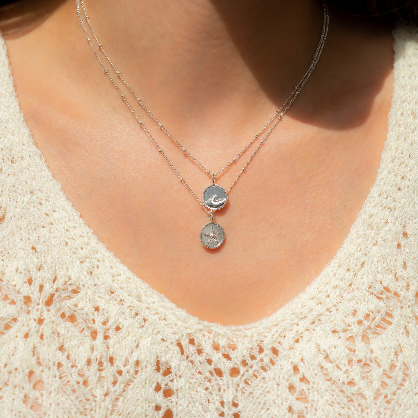 A person wearing a cream-colored crocheted shirt and two silver necklaces with circular pendants.