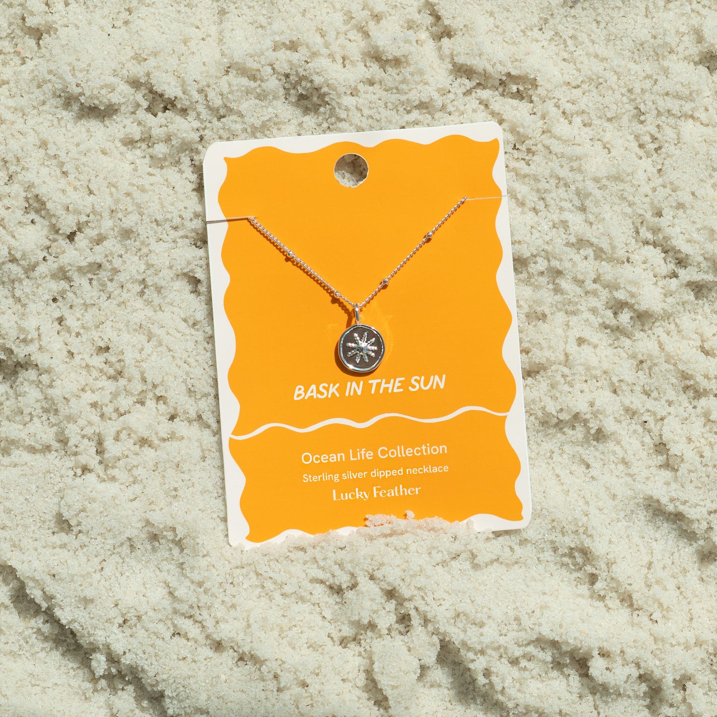 A sterling silver 'Lucky Feather' necklace on its orange display card rests on light sand.