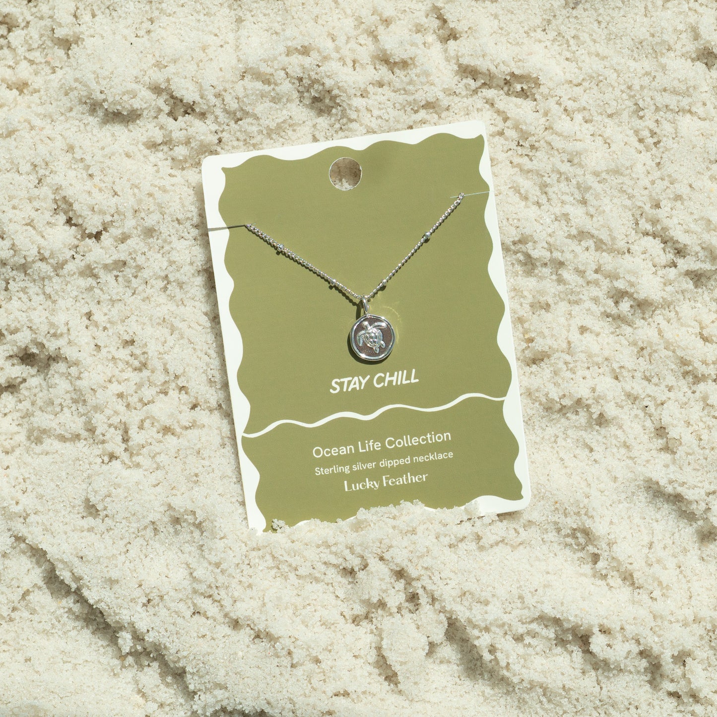A silver turtle pendant necklace on its packaging rests on sand.