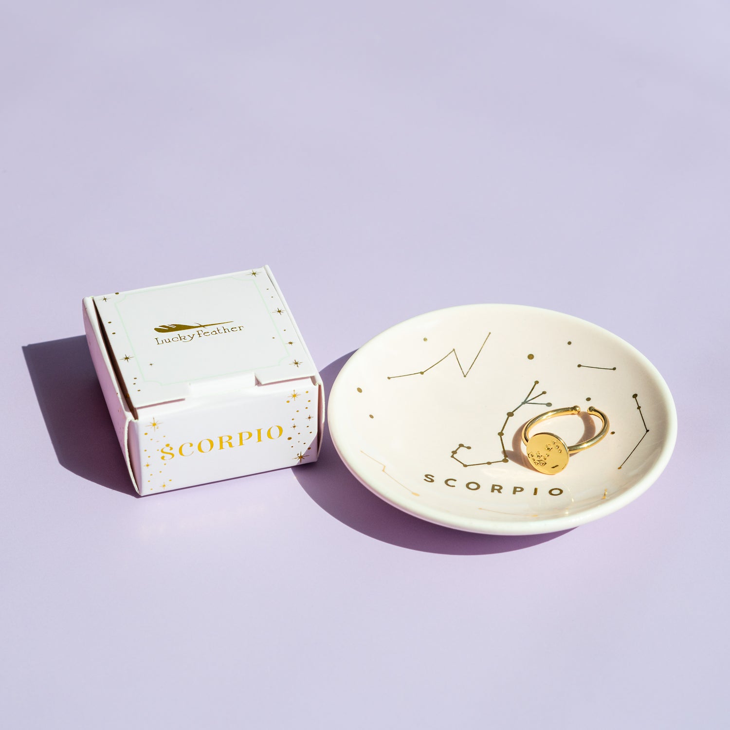 A 'Lucky Feather' box labeled 'Scorpio' is next to a small dish with the Scorpio constellation and a gold ring on a lavender background.