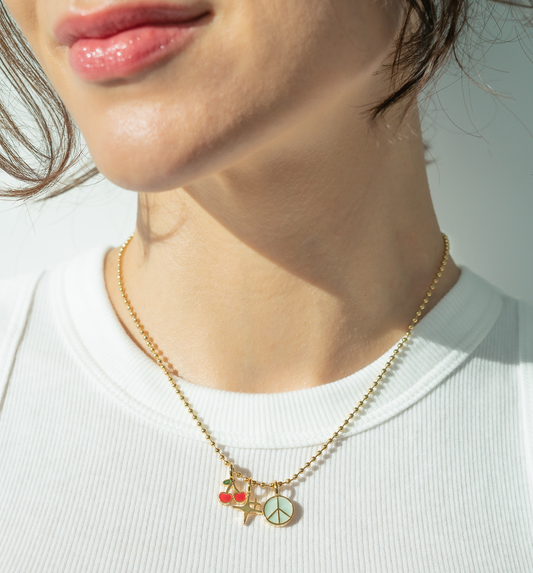A woman wears a gold ball chain necklace with cherry, star, and peace sign charms.