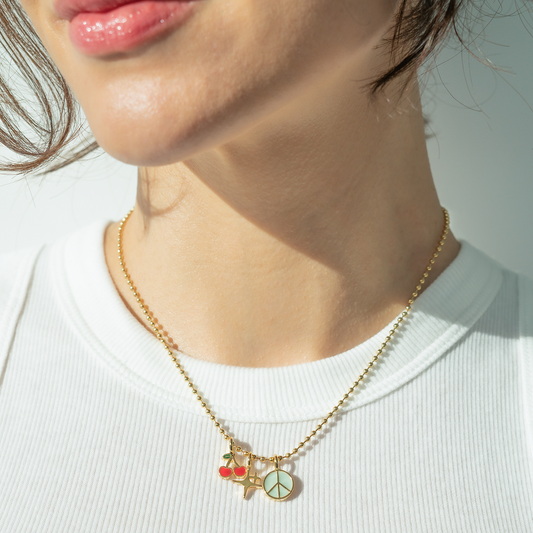 A woman wears a gold ball chain necklace with cherry, star, and peace sign charms.