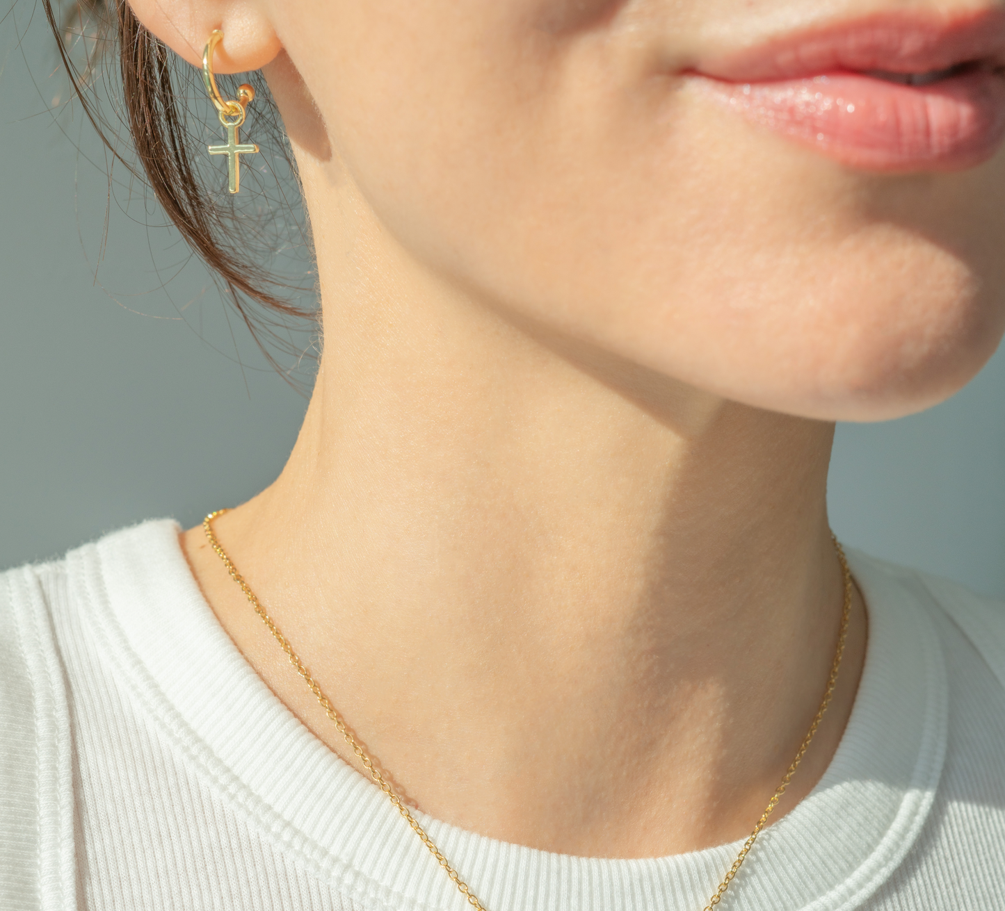 A close-up of a person's face and neck, showing a gold hoop earring with a cross and a gold necklace.