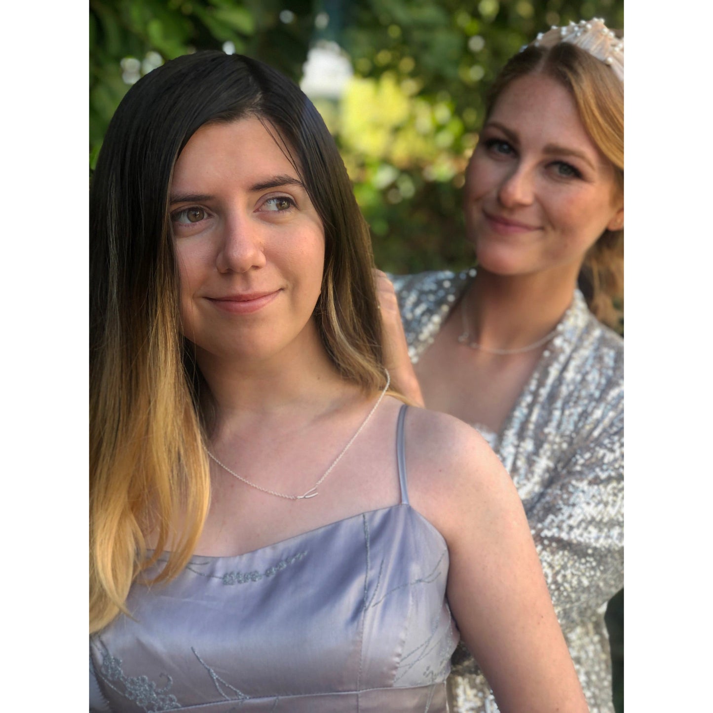 Two women are outdoors, one in a silver dress and necklace, and the other adjusting her hair, wearing a sequined shawl and pearl tiara.