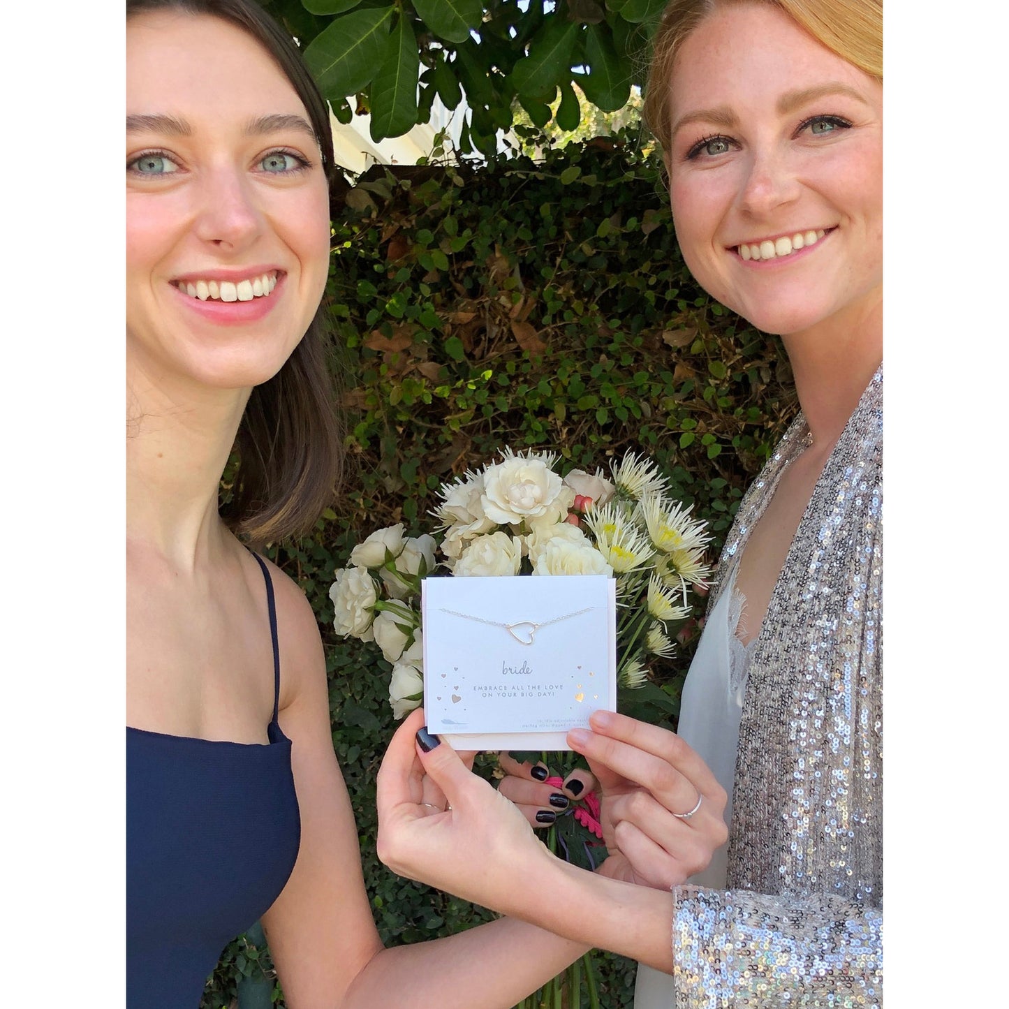 Two smiling women hold a 'bride' necklace in front of a bouquet of flowers and a green leafy background.