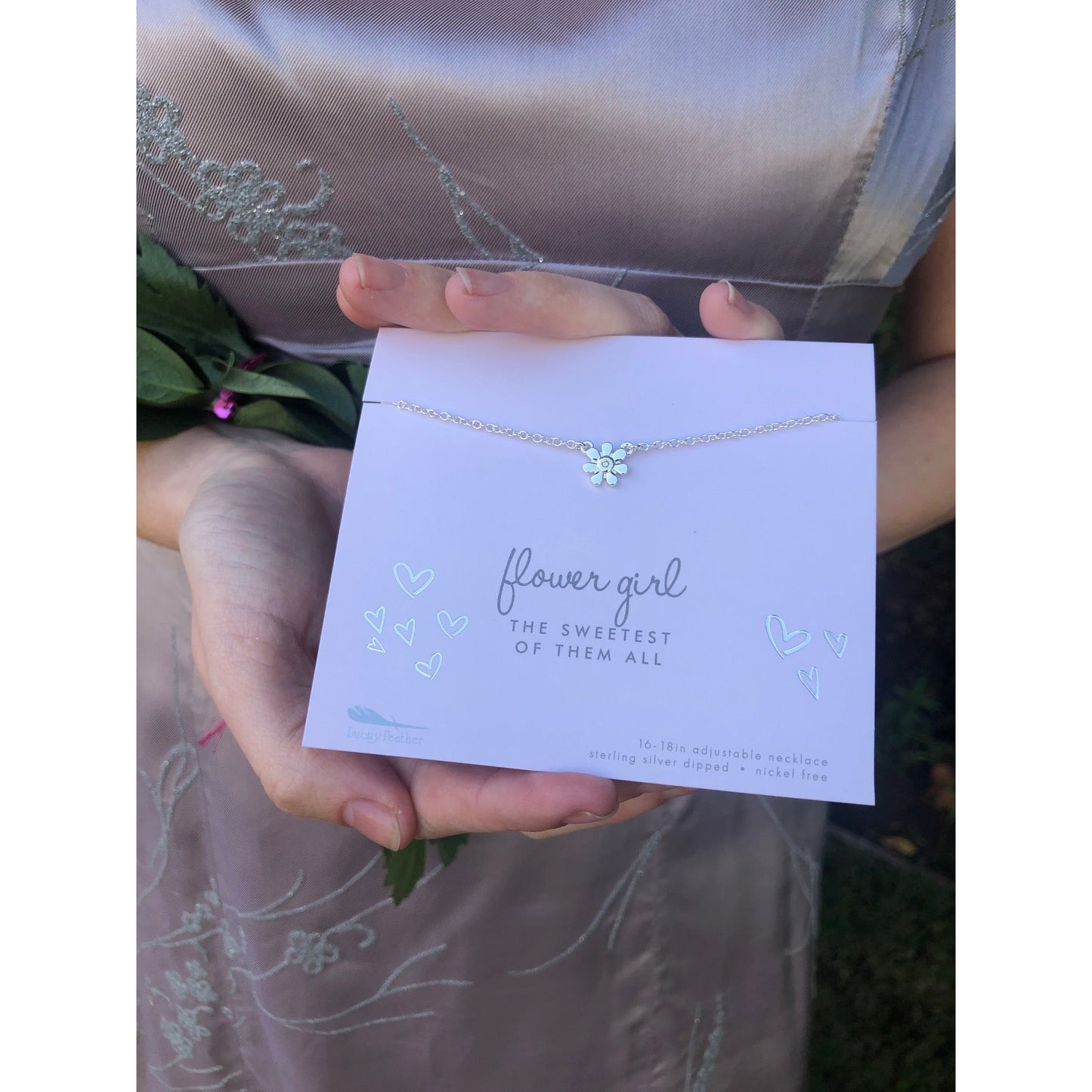 A person holds a 'flower girl' necklace with a flower charm on a card that reads 'flower girl, the sweetest of them all.'