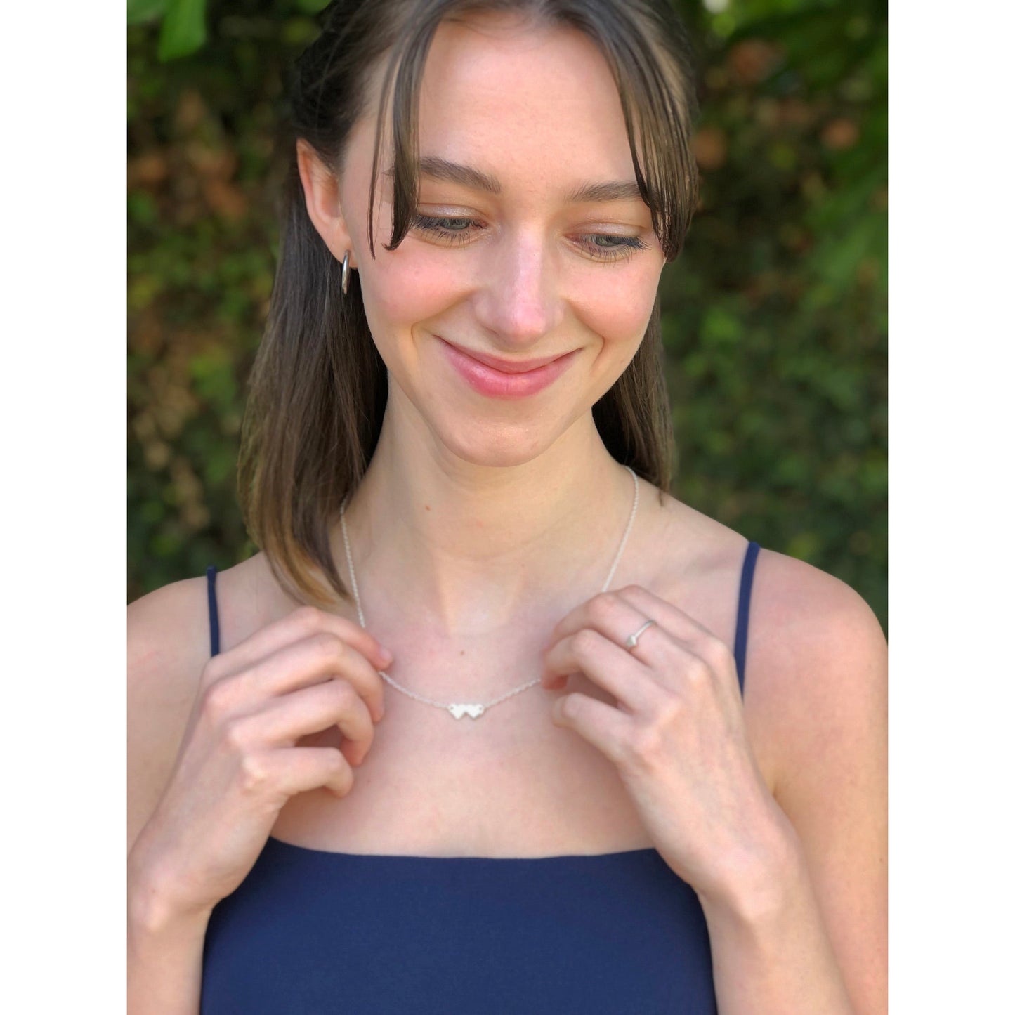 A smiling young woman with bangs is wearing a silver necklace with two connected heart pendants and a ring, standing in front of green foliage.
