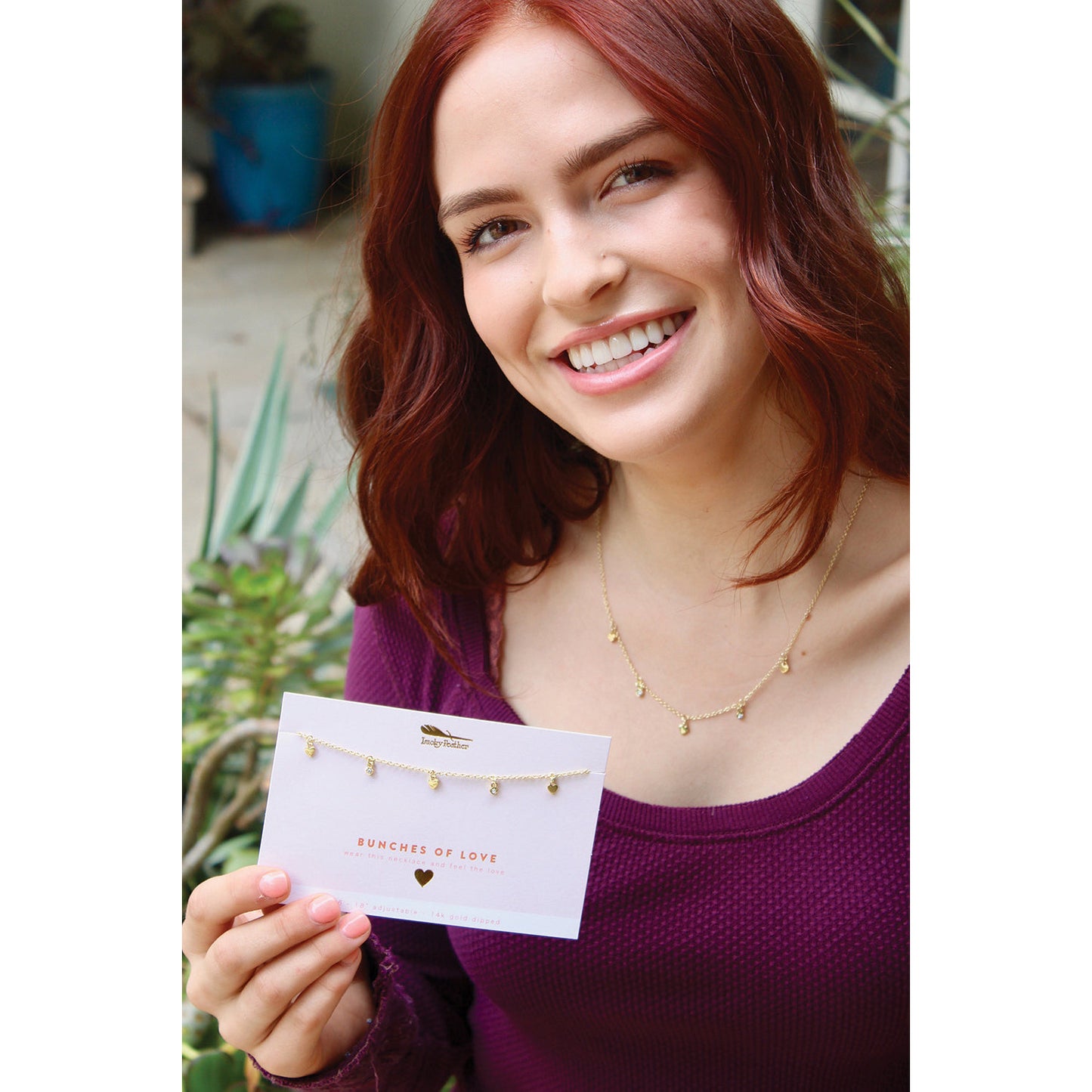 A smiling woman with red hair holds up a 'Bunches of Love' necklace.