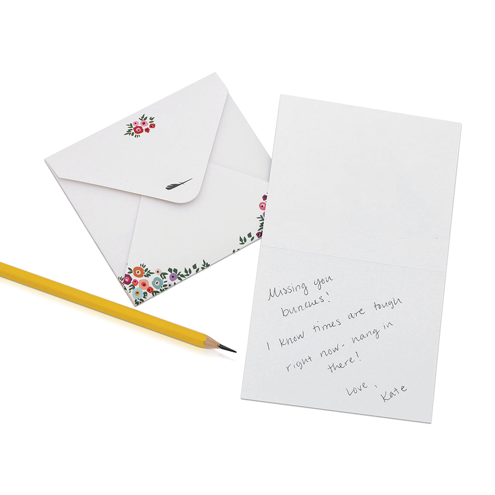 A floral-themed envelope, a greeting card with a handwritten message, and a yellow pencil lay on a white surface.