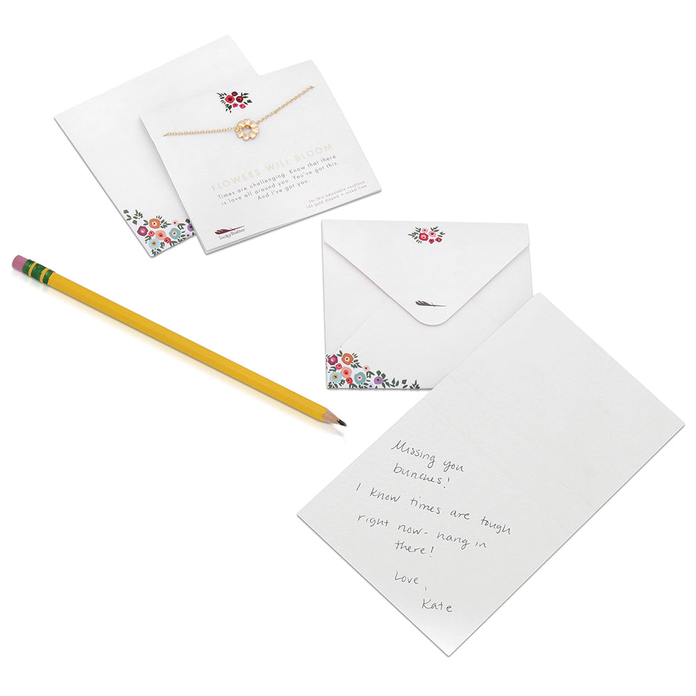 A gold necklace and handmade card with a handwritten message rest near an envelope and a yellow pencil.