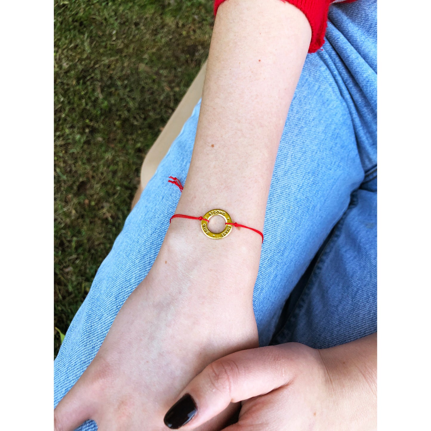 An arm wearing a red string bracelet with a gold ring charm rests on blue jeans.