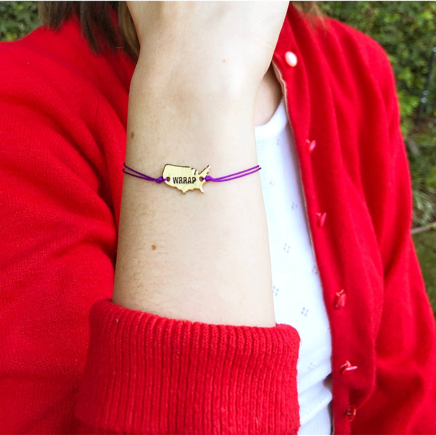 Person's wrist wearing a gold-colored bracelet with a United States shape and the word 'WRRAP' on a purple string, with a red cardigan sleeve visible.