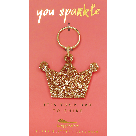 A glittery gold crown keychain is displayed on a pink card with gold lettering.