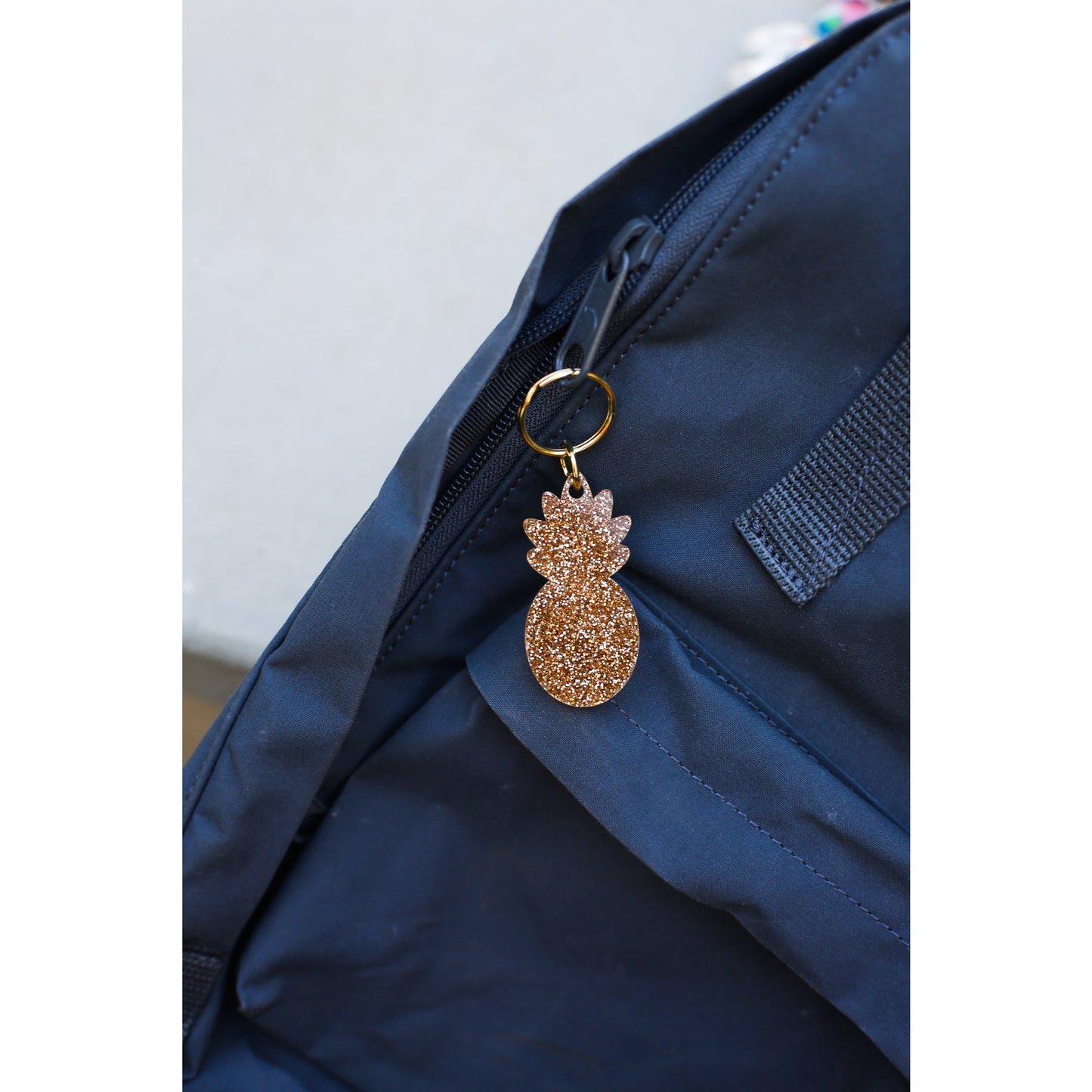 A gold glitter pineapple keychain hangs from the zipper of a navy blue backpack.