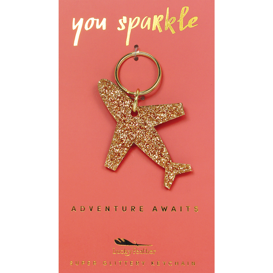 A gold glitter airplane keychain on a pink card with gold text that reads 'you sparkle' and 'adventure awaits.'