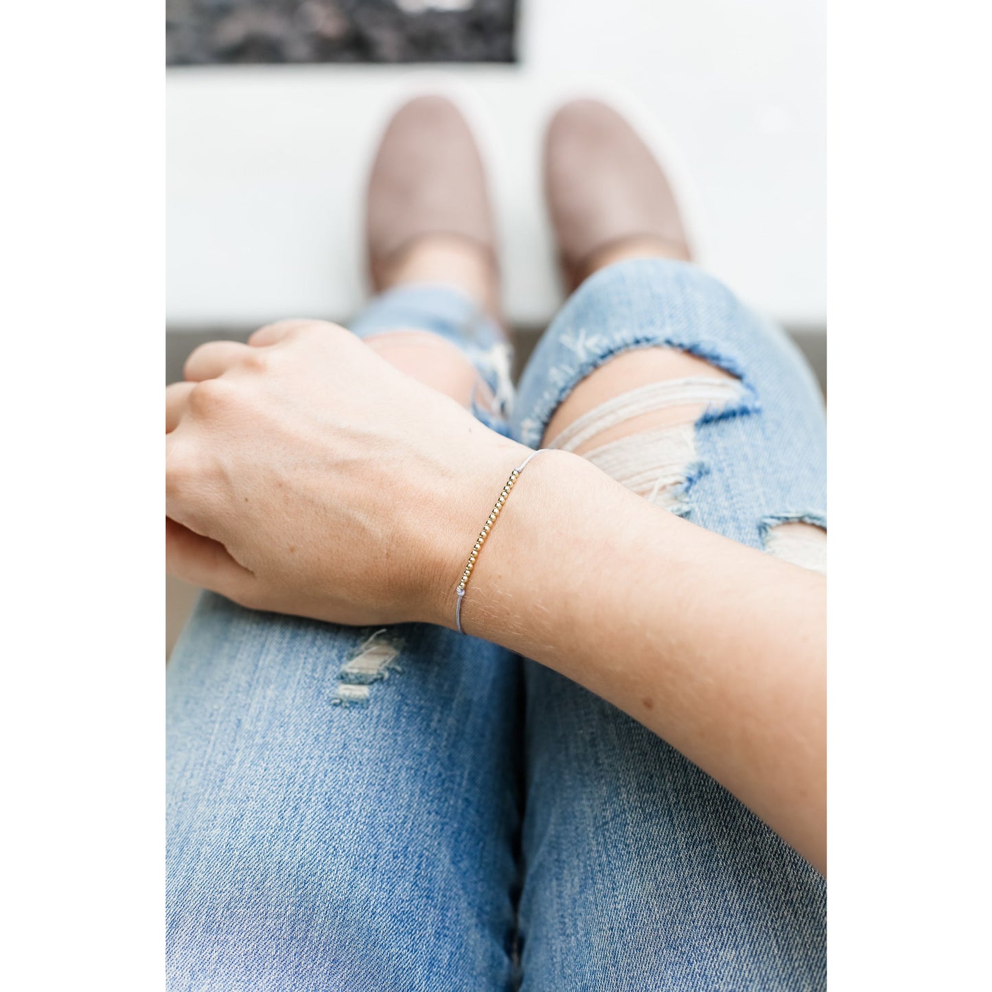 A person wearing ripped jeans and tan slip-on shoes has a gold beaded bracelet on their wrist.