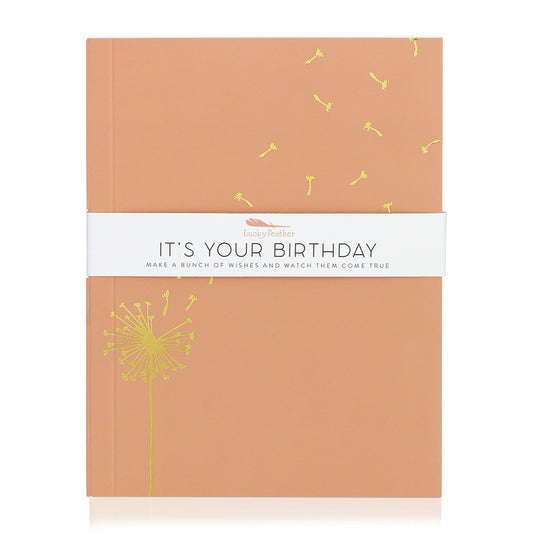 An orange journal wrapped in a white band that reads 'Lucky Feather IT'S YOUR BIRTHDAY MAKE A BUNCH OF WISHES AND WATCH THEM COME TRUE' is featured on a white background.