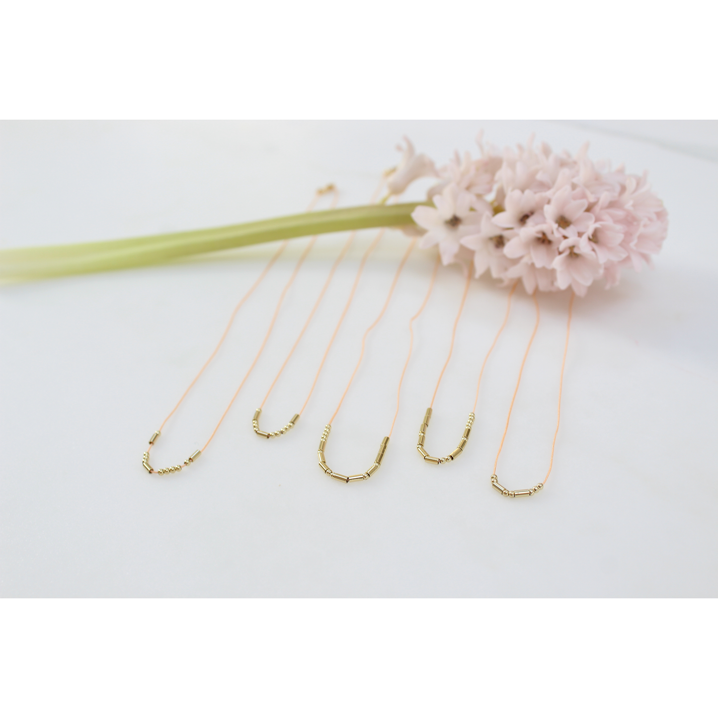 Several necklaces with gold-colored cylindrical and spherical beads are arranged next to a pink hyacinth flower on a white surface.