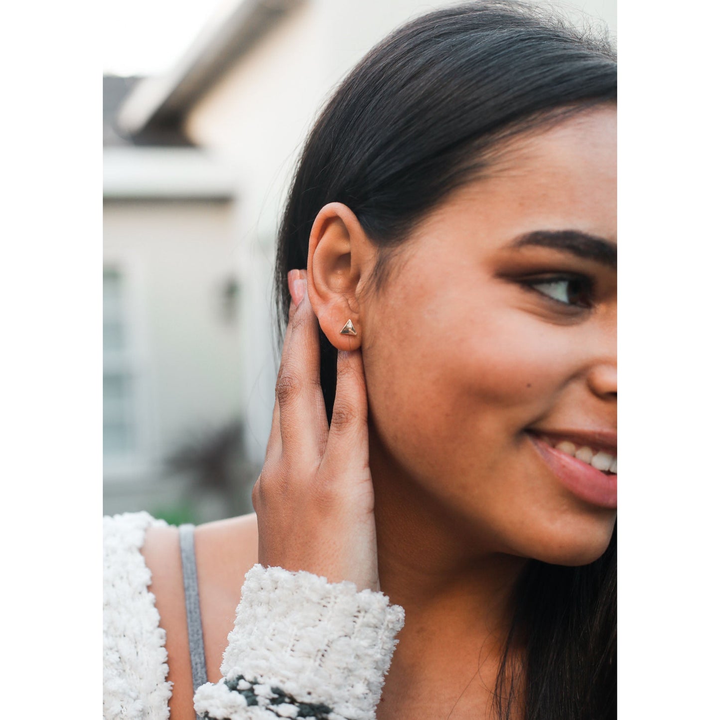 A smiling woman with dark hair is wearing a triangular stud earring and has her hand near her ear.