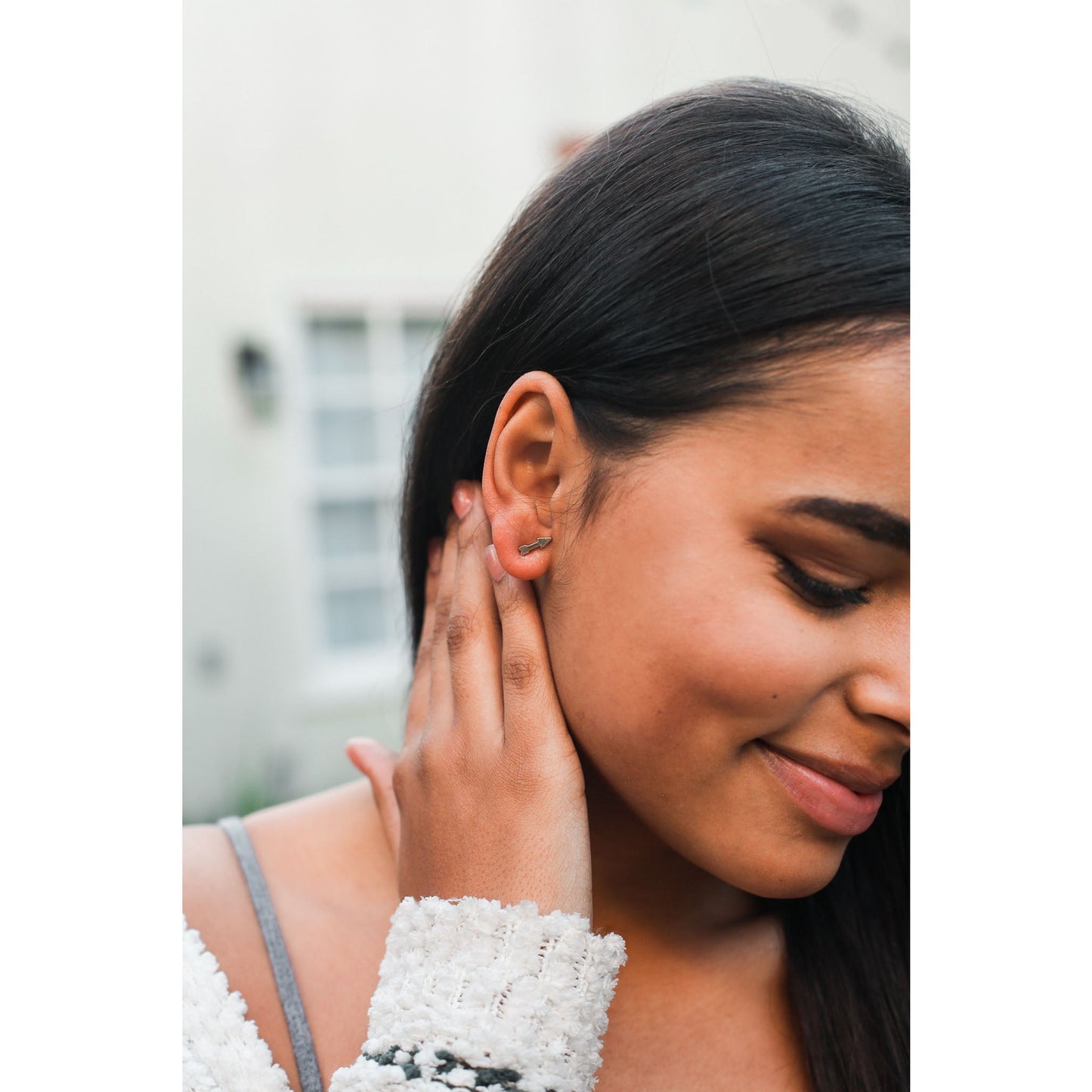 A woman with dark hair and a fair complexion wearing an arrow earring touches the back of her neck.