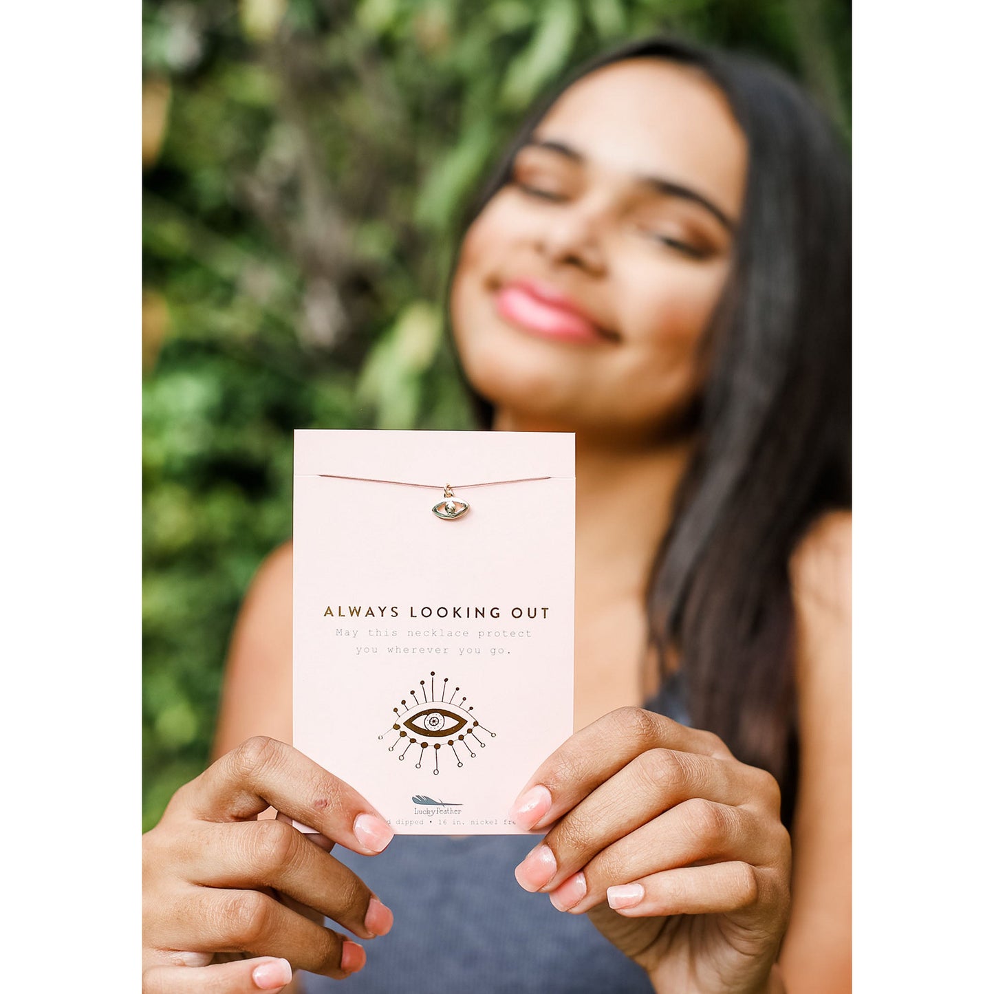 A woman holding an 'Always Looking Out' necklace against an out-of-focus green background.
