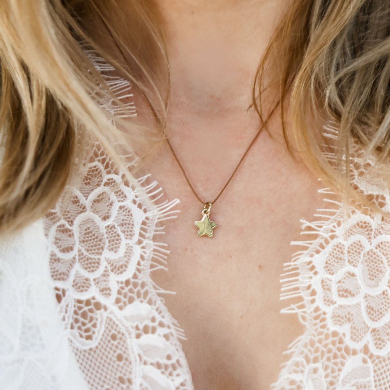 A woman wears a delicate golden star pendant on a thin brown cord necklace, against a background of white lace and blonde hair.