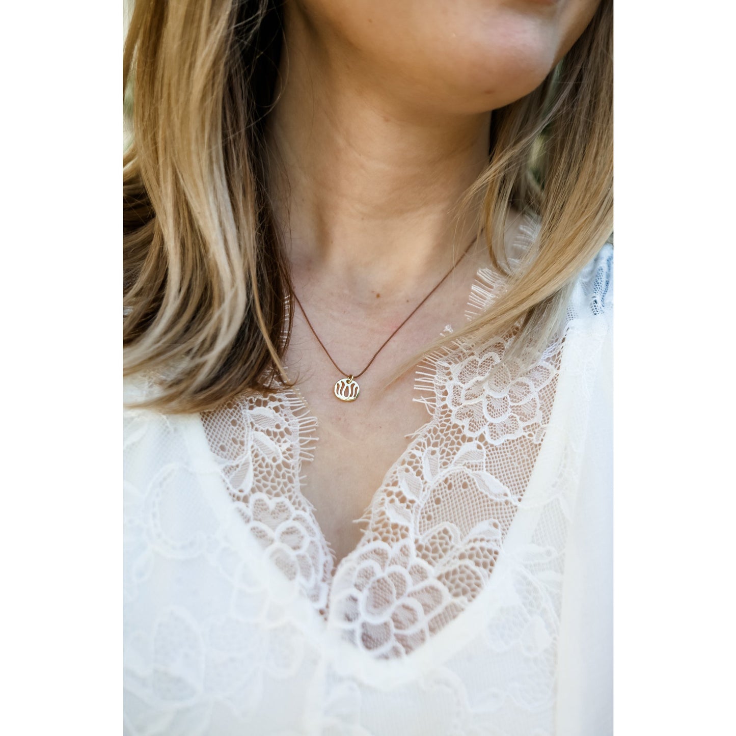 A person wears a gold pendant on a brown cord necklace with a white lace top.