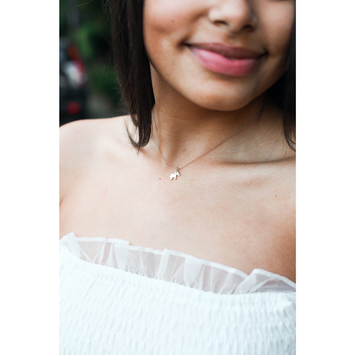 A smiling person wears a white strapless dress and a delicate necklace with an elephant pendant.