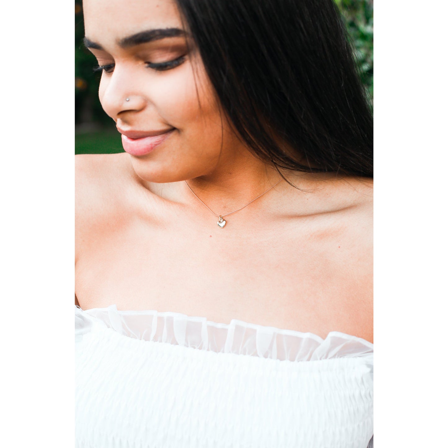 A woman with dark hair and a nose piercing wears a white top and a heart pendant necklace.