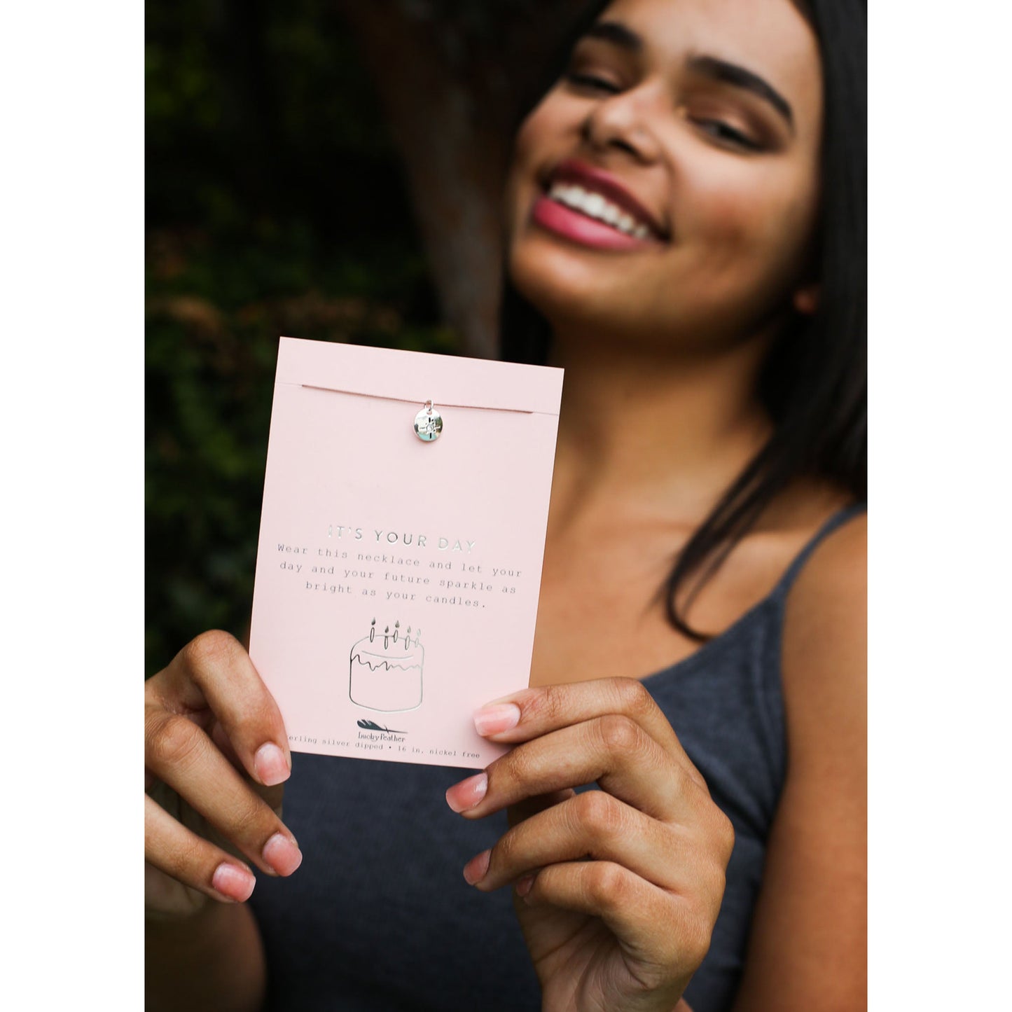 A smiling woman holds a card with a necklace attached, reading 'It's Your Day'.